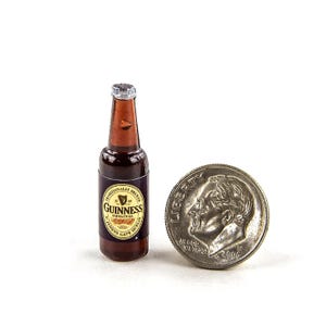 May include: A miniature Guinness beer bottle with a silver cap and a label that reads "Guinness Original". A coin is next to the bottle for scale. The bottle is brown and black.
