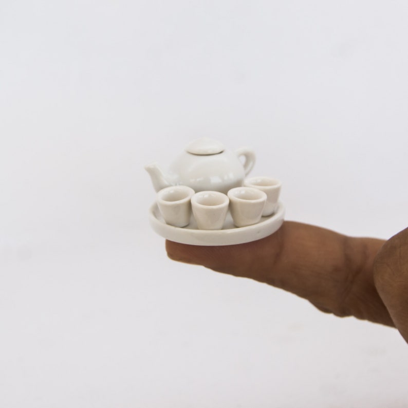 May include: A miniature white ceramic tea set, including a teapot with a lid, four small cups, and a round serving tray. The set is held in a person's hand against a white background. The tea set is a delicate and charming item.