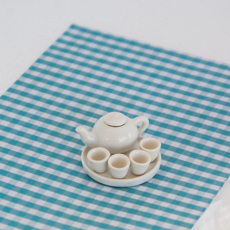 May include: A miniature white ceramic tea set, including a teapot with a lid and four small teacups, arranged on a round tray. The set is placed on a blue and white checkered surface, creating a charming and delicate aesthetic.