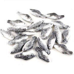 以下が含まれることがあります： 小さなセラミック製の魚の置物のコレクション。各魚はグレーと白の色合いで塗装されており、詳細な鱗とひれがあります。白い表面に配置された置物です。