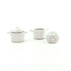 May include: Three white ceramic pots with lids. The pots are small and have rounded handles. The lids have a rounded knob on top.