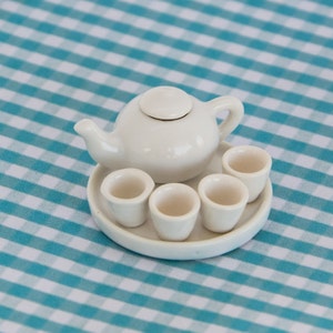 May include: A miniature white ceramic tea set, including a teapot with a lid, a serving tray, and four small teacups. The set is displayed on a blue and white checkered tablecloth.