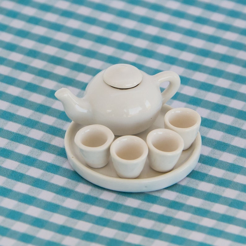 May include: A miniature white ceramic tea set, including a teapot with a lid, a serving tray, and four small teacups. The set is displayed on a blue and white checkered tablecloth.