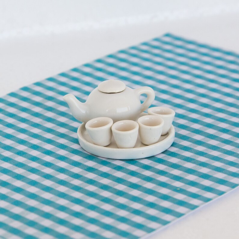 May include: A miniature white ceramic tea set, including a teapot and four cups on a small tray. The set is placed on a blue and white checkered surface, suggesting a playful or dollhouse theme. The teapot has a lid and a curved handle.