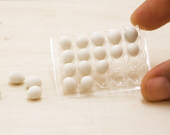 May include: A clear plastic egg carton filled with small, white eggs. The carton is held by a person's fingers. Several loose eggs are scattered on a light-colored wooden surface. The eggs appear to be miniature or decorative.