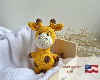 May include: Crochet pattern PDF for a cute yellow and brown giraffe amigurumi toy. The giraffe is sitting in a wooden crate with white fabric.