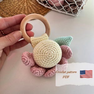 May include: A crocheted baby rattle with a wooden ring. The rattle is made of soft yarn in shades of pink, yellow, and green. The crocheted flower has a center circle and petals. The rattle is attached to the wooden ring with a crocheted strap.