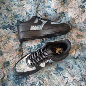 May include: A pair of black Nike Air Force 1 sneakers with a floral tapestry pattern. The sneakers have black laces and a white swoosh logo.
