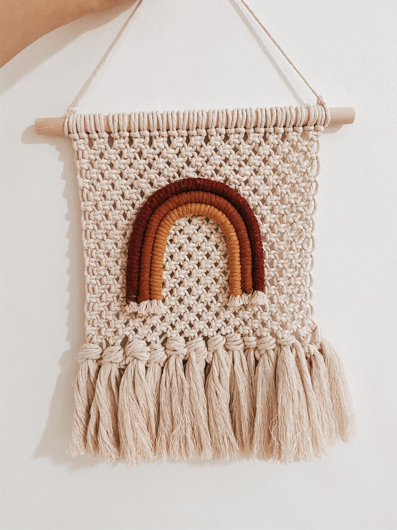 Pode incluir: Uma tape&ccedil;aria de macram&eacute; bege com um design de arco-&iacute;ris em tons de marrom, laranja e vermelho. A tape&ccedil;aria tem uma barra de madeira na parte superior e uma franja na parte inferior.