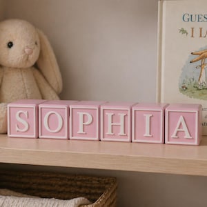 May include: A set of pink wooden blocks spelling "SOPHIA" in white capital letters. A soft toy rabbit and a children's book, "Guess How Much I Love You", are visible, creating a nursery or children's room setting.