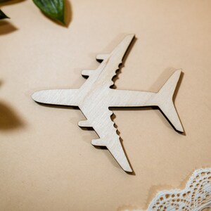 May include: A wooden airplane cutout on a beige background with green leaves and white lace in the corners.