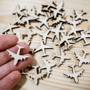 May include: A collection of wooden airplane cutouts scattered on a light wood surface. One cutout is being held by a hand.