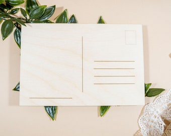 May include: A blank wooden postcard with a space for a stamp and lines for an address and message. The postcard is surrounded by green leaves and white lace.