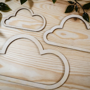 May include: Three wooden cloud shapes of varying sizes, arranged on a light wood surface. The clouds are cut out and have a smooth, natural finish.