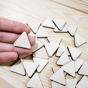 May include: A hand holding a small, plain wooden triangle.  Surrounding the hand are many more wooden triangles scattered on a light wood surface.