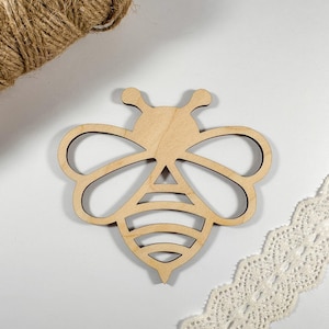 May include: A wooden bee-shaped cutout with a detailed design. The cutout is light brown and sits on a white surface, accompanied by a roll of twine and a piece of white lace trim. The bee has a detailed body.