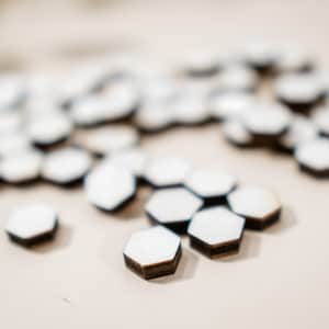 Puede incluir: Primer plano de un grupo de azulejos hexagonales blancos con bordes de color marrón oscuro. Los azulejos están dispersos sobre una superficie de color beige claro.