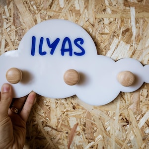 May include: A white cloud-shaped wall hook with the name "ILYAS" in blue lettering. It features three wooden knobs for hanging items. The hook is held against a wooden background.