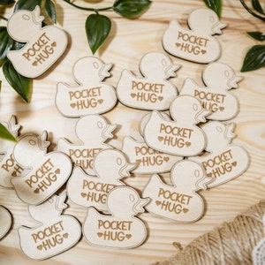 Op de afbeelding: Een verzameling houten eendvormige ornamenten met de tekst "Pocket Hug" erop gegraveerd. De ornamenten liggen verspreid op een houten oppervlak.