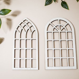 May include: Two white wooden arched window frames with a grid pattern. The frames are on a light beige background.