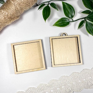 Può includere: Due cornici quadrate in legno non rifinite con venatura chiara, una con un piccolo anello nella parte superiore. Un rotolo di spago e foglie verdi sullo sfondo. Rifinitura in pizzo bianco in basso.