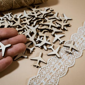 May include: A close-up of a pile of wooden airplane cutouts. The cutouts are a light brown color and are scattered on a beige surface. A hand is holding one of the cutouts.