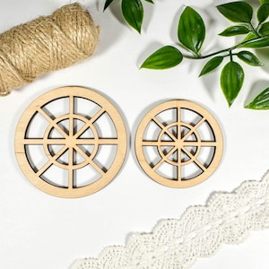 May include: Two circular wooden decorative pieces with a geometric design, like ship's wheels. A roll of twine and white lace are also present. Green leaves add a natural touch. The pieces are made of light-coloured wood. The diameter of the larger piece is approximately 10 cm.