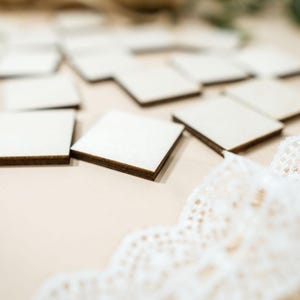 Op de afbeelding: Een verzameling kleine, vierkante houten tegels met donkerbruine randen, verspreid over een lichtbeige oppervlak. Een delicate witte kanten lint is gedeeltelijk zichtbaar. De tegels lijken blanco en klaar voor personalisatie.