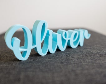 Könnte beinhalten: Ein hellblaues Holz-Namensschild mit der Aufschrift "Oliver" in kursiv.