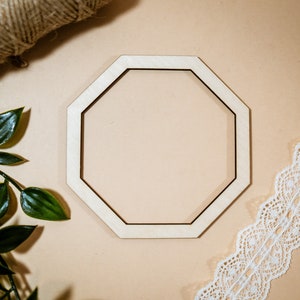 May include: A wooden octagon-shaped frame with a light brown finish. The frame is on a beige background with a piece of white lace and green foliage.