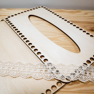 Op de afbeelding: Twee onafgewerkte houten stukken voor een DIY geweven tas project. De stukken zijn gemaakt van licht hout en hebben gaten rond de randen. Een witte kantversiering is op de stukken geplaatst.