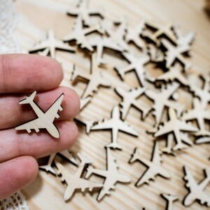 May include: A close-up of a hand holding a small wooden airplane cutout. The cutout is surrounded by many other wooden airplane cutouts, all scattered on a light brown wooden surface.