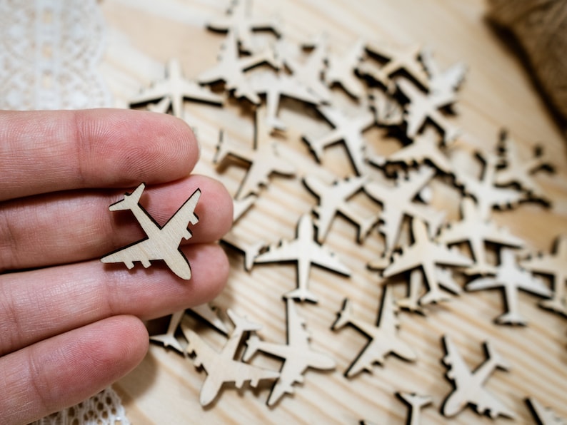 May include: A close-up of a hand holding a small wooden airplane cutout. The cutout is surrounded by many other wooden airplane cutouts, all scattered on a light brown wooden surface.