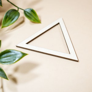 Puede incluir: Un marco de madera blanco en forma de triángulo sobre un fondo beige con hojas verdes en primer plano.