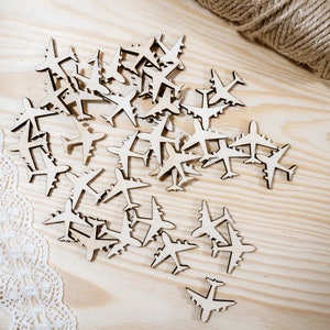 May include: A large pile of wooden airplane cutouts on a light brown wooden surface. The cutouts are all the same size and shape, and they are arranged in a random pattern. There is a piece of white lace on the left side of the image.