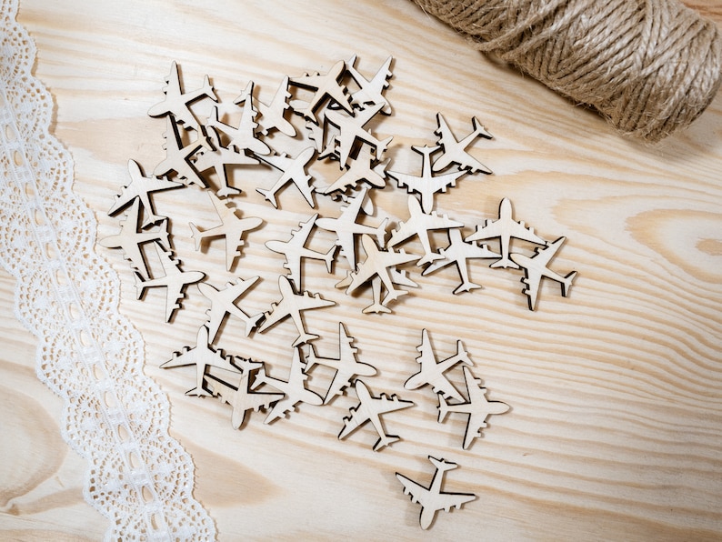 May include: A large pile of wooden airplane cutouts on a light brown wooden surface. The cutouts are all the same size and shape, and they are arranged in a random pattern. There is a piece of white lace on the left side of the image.