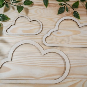 May include: Three wooden cloud shapes of varying sizes, arranged on a light wood surface. The clouds are cut out of the wood, creating a frame-like effect.