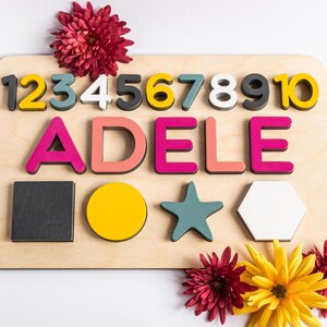 May include: Wooden board with colorful numbers 1 through 10, the name "ADELE" spelled out in pink and red letters, and geometric shapes including a square, circle, star, and hexagon. The board is surrounded by red and yellow flowers.