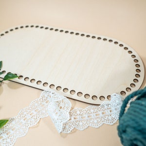 Op de afbeelding: Een ovale houten weefgetouw met gaten rond de rand, een spoel turquoise garen en een witte kantversiering.