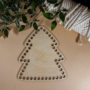 Könnte beinhalten: Ein hölzerner Weihnachtsbaum-Webrahmen mit Löchern an den Rändern zum Weben von Garn. Der Webrahmen ist hellbraun und liegt auf einer beigefarbenen Oberfläche.
