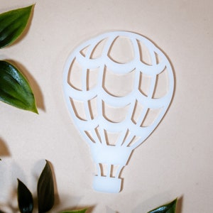 Pode incluir: Corte de papel branco de um balão de ar quente com um design detalhado. O balão está sobre um fundo bege claro.