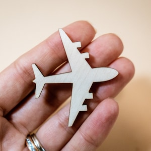 Puede incluir: Un avión de madera recortado sostenido en una mano. El avión es blanco y tiene un diseño simple.