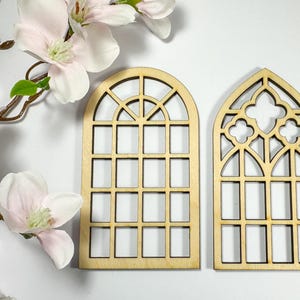 Könnte beinhalten: Zwei dekorative Fenster-Ausschnitte aus Holz. Eines ist gewölbt mit einem Gittermuster, das andere spitz mit einem Vierpass-Design. Die Fenster sind aus hellem Holz gefertigt und vor einem weißen Hintergrund mit rosa Blüten platziert.