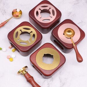 May include: Four wooden candle warmers with brass accents. Each warmer has a square base and a round top with a hole in the center. The warmers are in different colors, including brown, gold, and copper. There are also two spoons and a wax seal stamp.