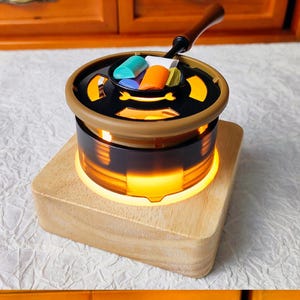 May include: A small, round, black and gold wax melter with a wooden base. The melter has a clear glass bowl with a light inside, and a metal top with a handle. There are four colorful wax melts in the bowl.