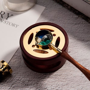 May include: A gold-colored wax seal melting spoon with a wooden handle sits in a wooden base with a gold-colored metal top. The spoon is filled with colorful wax beads.
