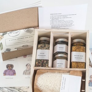 May include: A wooden box set containing jars of spices, a bag of rice, and recipe cards. The jars are labeled "Onion Seasoning," "Pepper," "Chicken Salt," and "Yellow Rice Seasoning." The recipe cards feature illustrations and text.