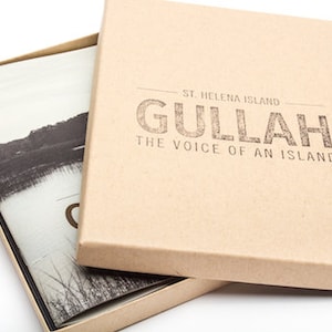 Może przedstawiać: Brązowe pudełko kartonowe z białą książką w środku. Na przodzie pudełka znajduje się napis "St. Helena Island Gullah The Voice of an Island".