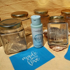 Peut inclure: Un flacon bleu de crème à graver le verre avec les mots "Magic Glass" et "Cadence" sur l'étiquette.  Plusieurs bocaux en verre avec des couvercles dorés et les mots "made with love" gravés dessus.  Un pochoir bleu avec les mots "made with love" est sur la table.