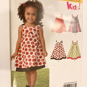 Könnte beinhalten: Ein New Look Schnittmuster für Kinderkleider. Das Schnittmuster zeigt verschiedene Kleiderdesigns, darunter ein rot-weiß gepunktetes Kleid. Das Bild enthält den Text "NEW LOOK" und "Kids!"
